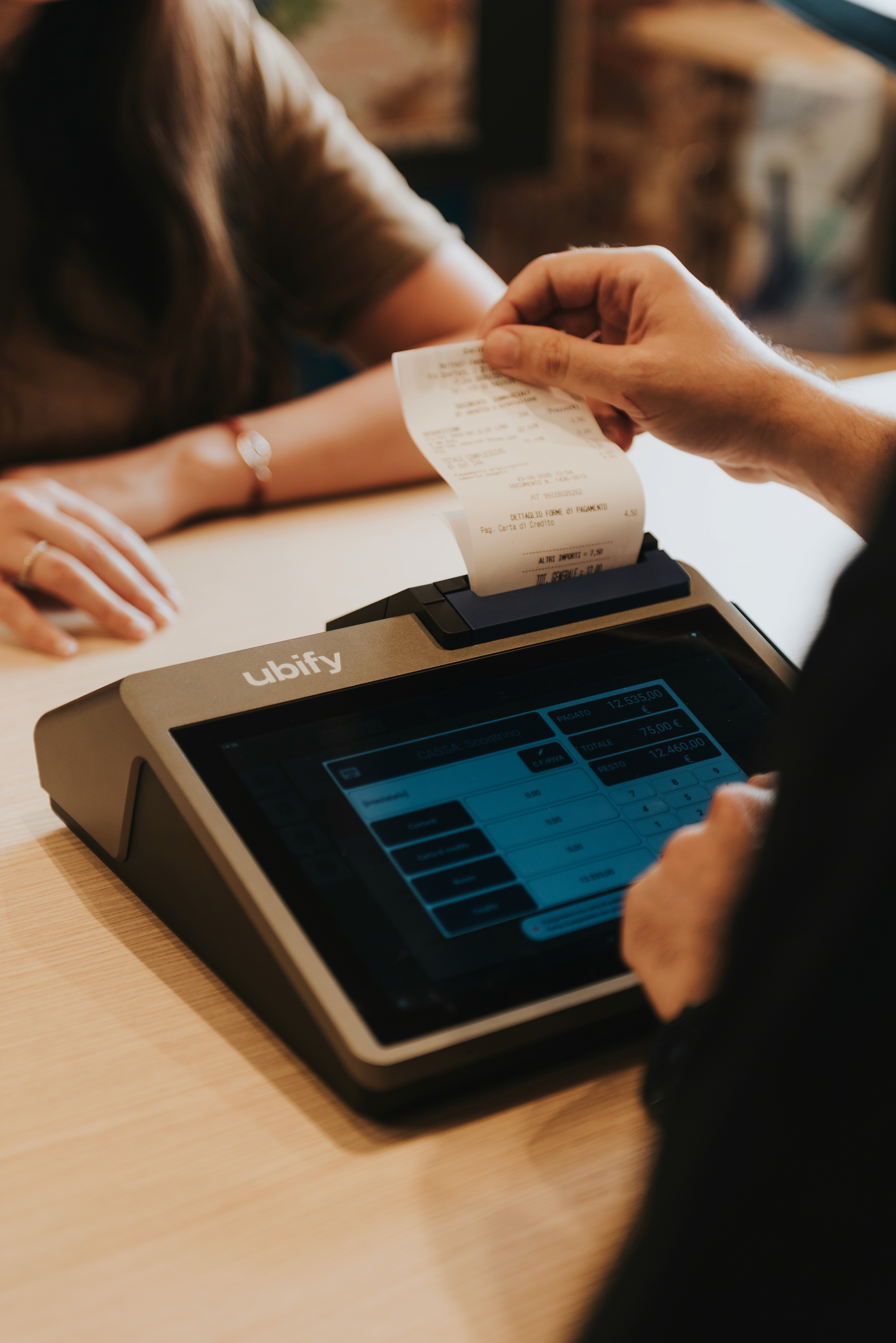 Cliente riceve uno scontrino stampato dalla Cassa Ubify, con interfaccia touchscreen attiva sul bancone.
