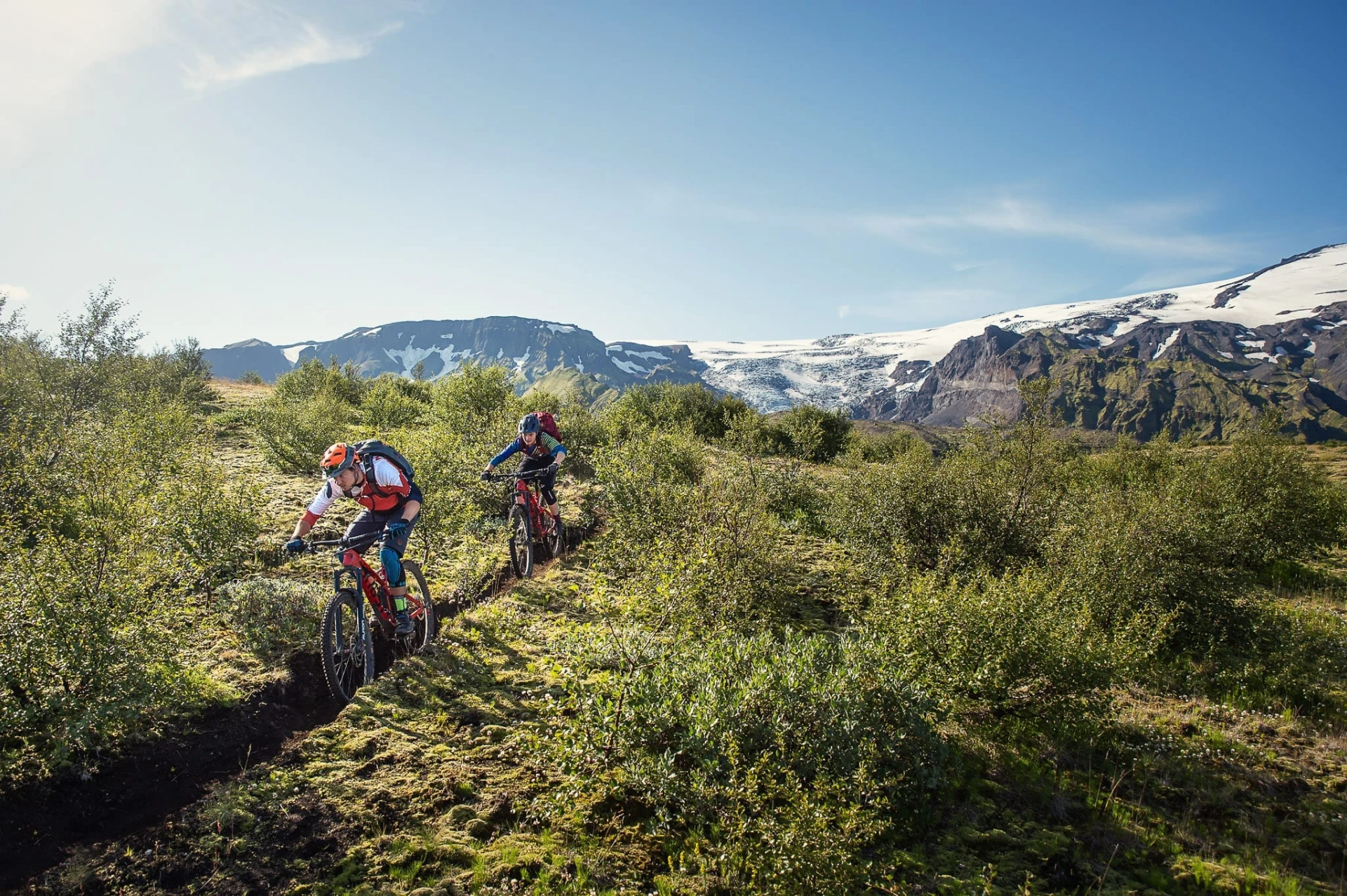 Two mountain bikers riding a narrow trail with a glacier and mountains behind.