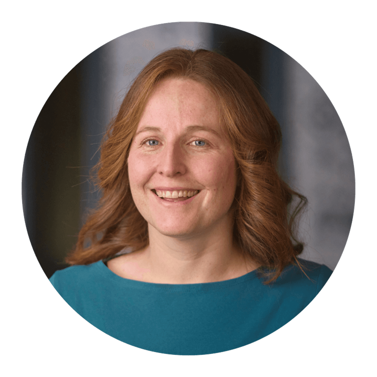 A circular studio portrait of a person with shoulder‑length auburn hair, wearing a teal top, set against a softly blurred grey backdrop.