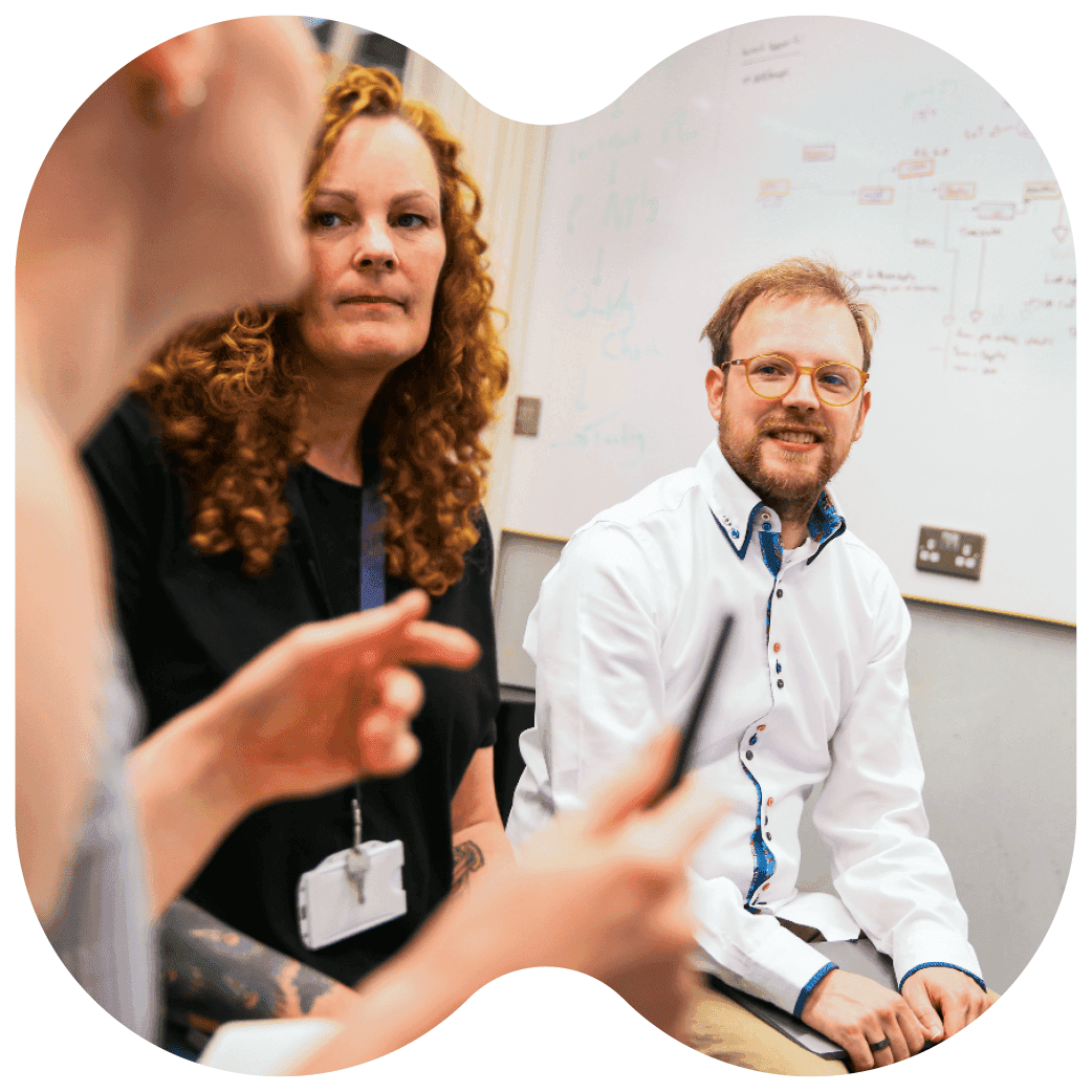 People in a meeting room engaged in discussion, with one person holding a pen and a whiteboard with diagrams in the background.