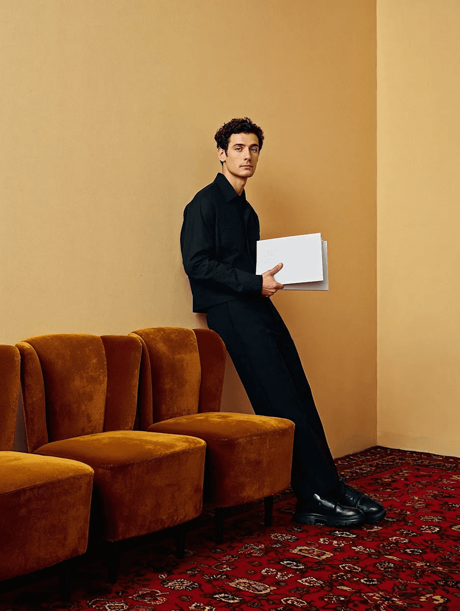 Man dressed in black leaning against a mustard-colored wall while holding white folders in a room with patterned carpet and velvet chairs.