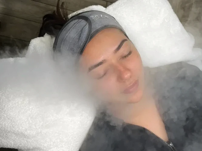 A person wearing a gray headband relaxes with eyes closed, experiencing a soothing facial steam treatment. They appear calm and content.