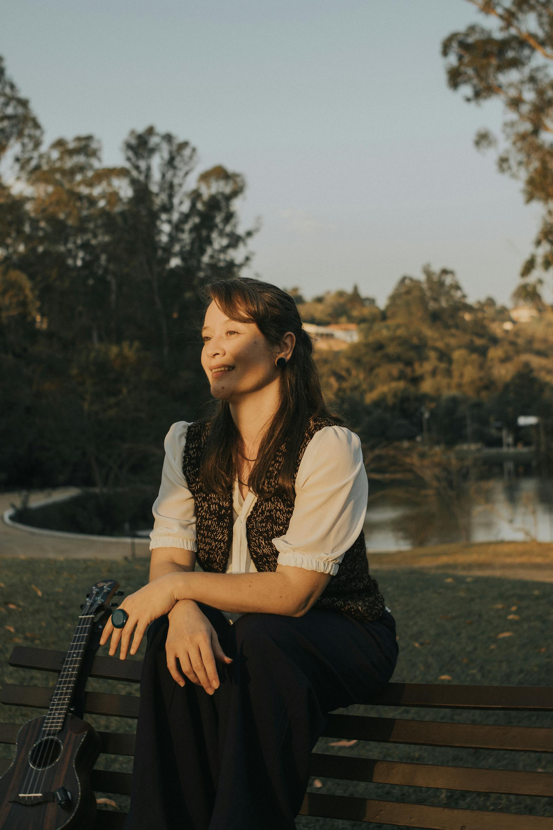 A woman in a white blouse and dark vest sits on a park bench, looking away and smiling. A ukulele rests next to her.