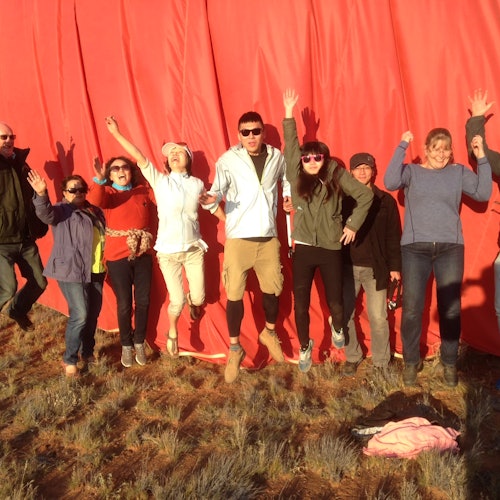 A group of eight people jumping and smiling in front of a large red fabric backdrop on a grassy field.