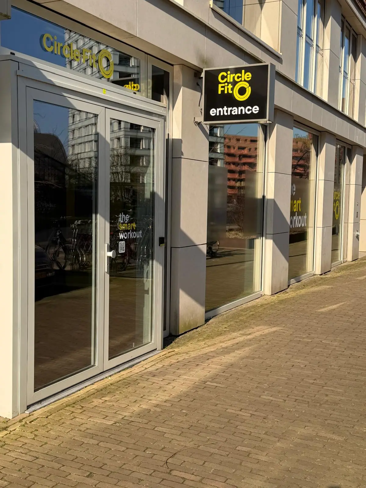 A street view of a Circle Fit hub with large glass doors and yellow banners, alongside a parked car.