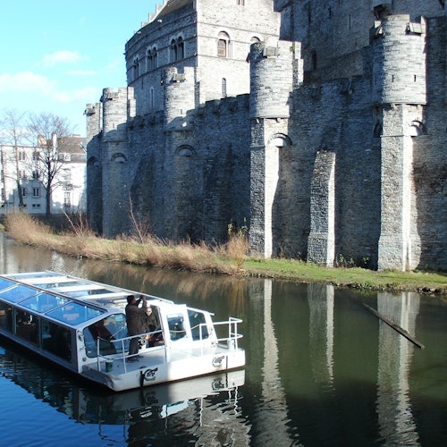 A tour boat floats near a castle with tall stone walls along a calm river under a clear blue sky.