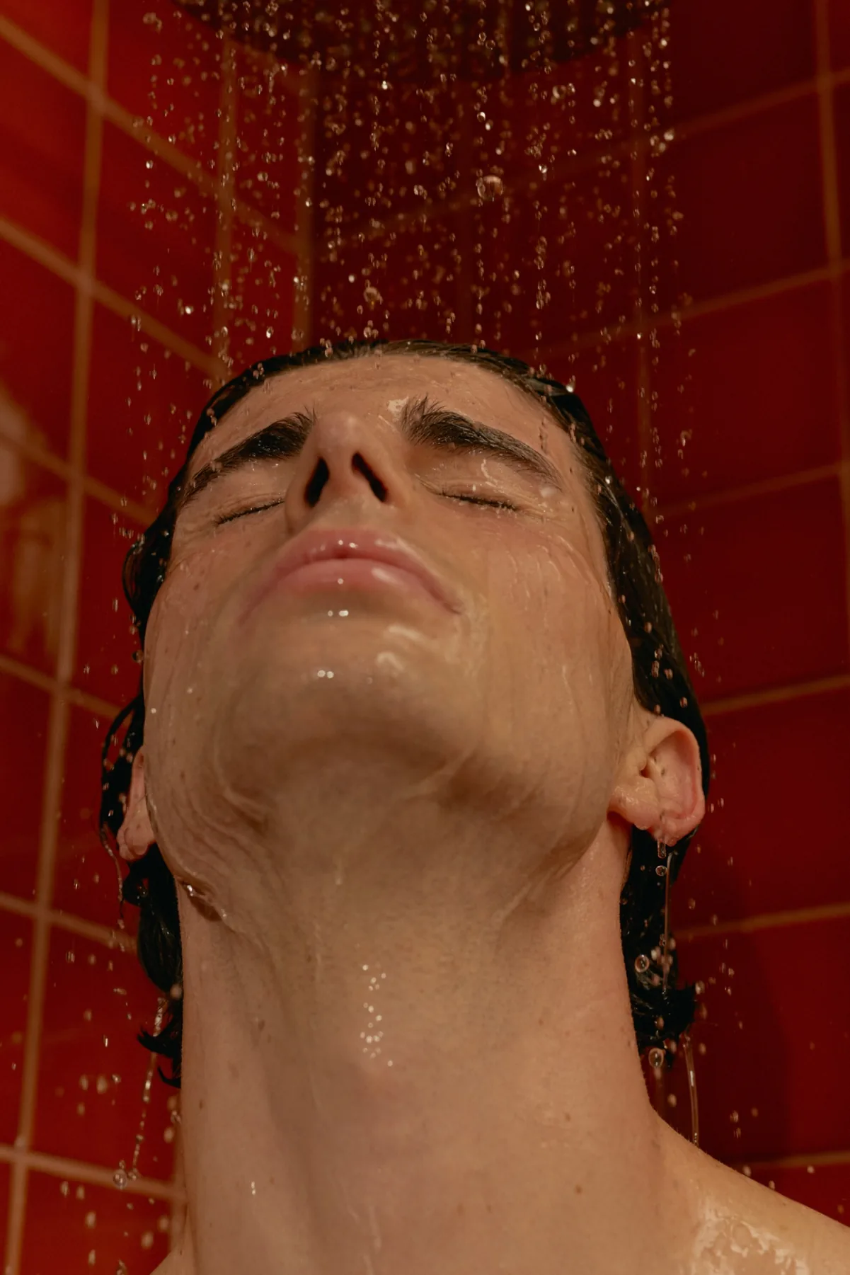 A man with his eyes closed under a shower against red tiles