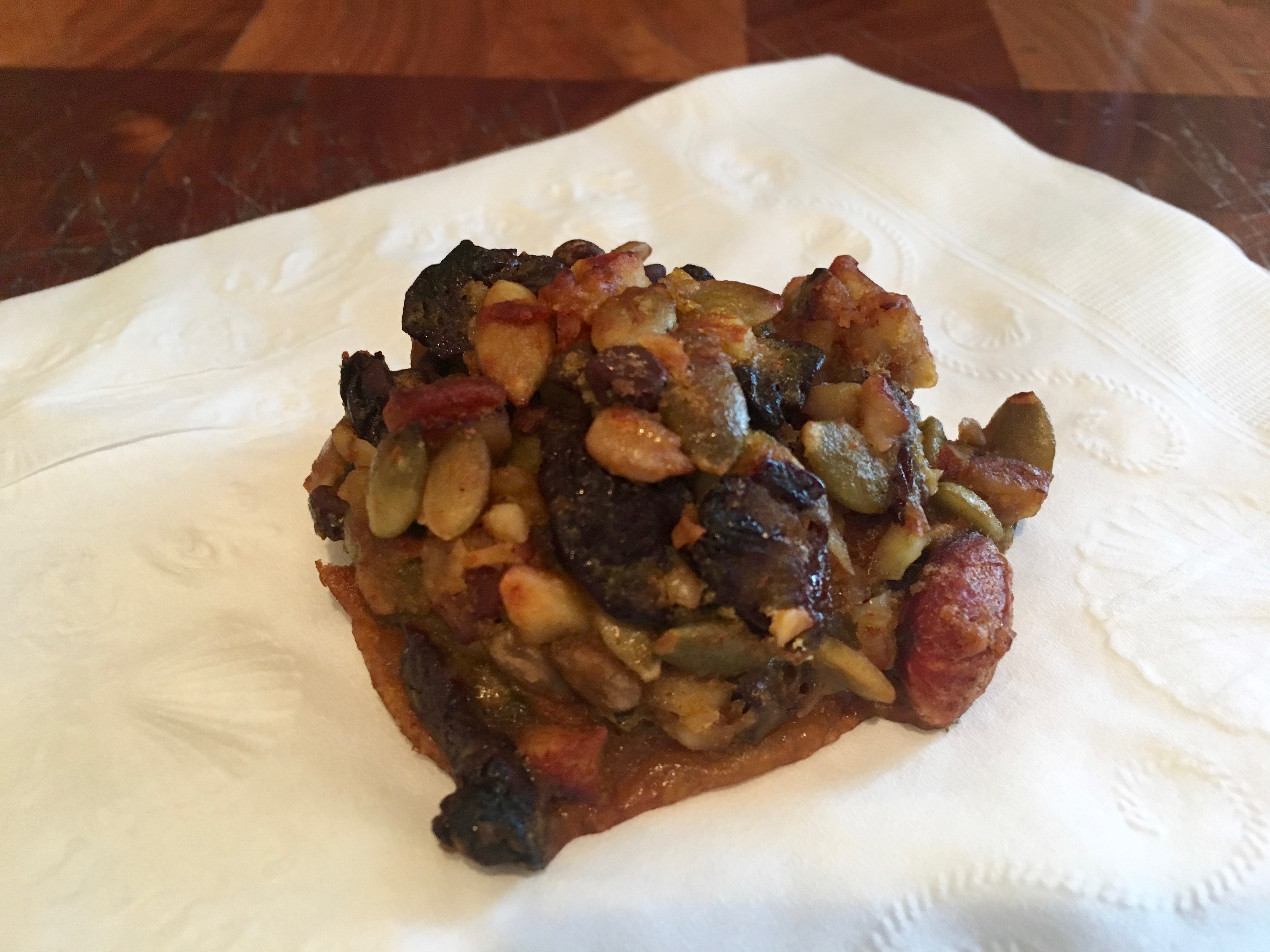 A flourless pumpkin power cookie resting on a white paper, featuring a mix of nuts and fruits, with a golden-brown crust.