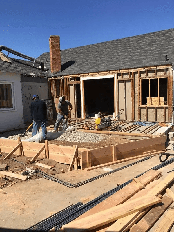 Construction workers spreading gravel inside wooden concrete forms during a home patio renovation.