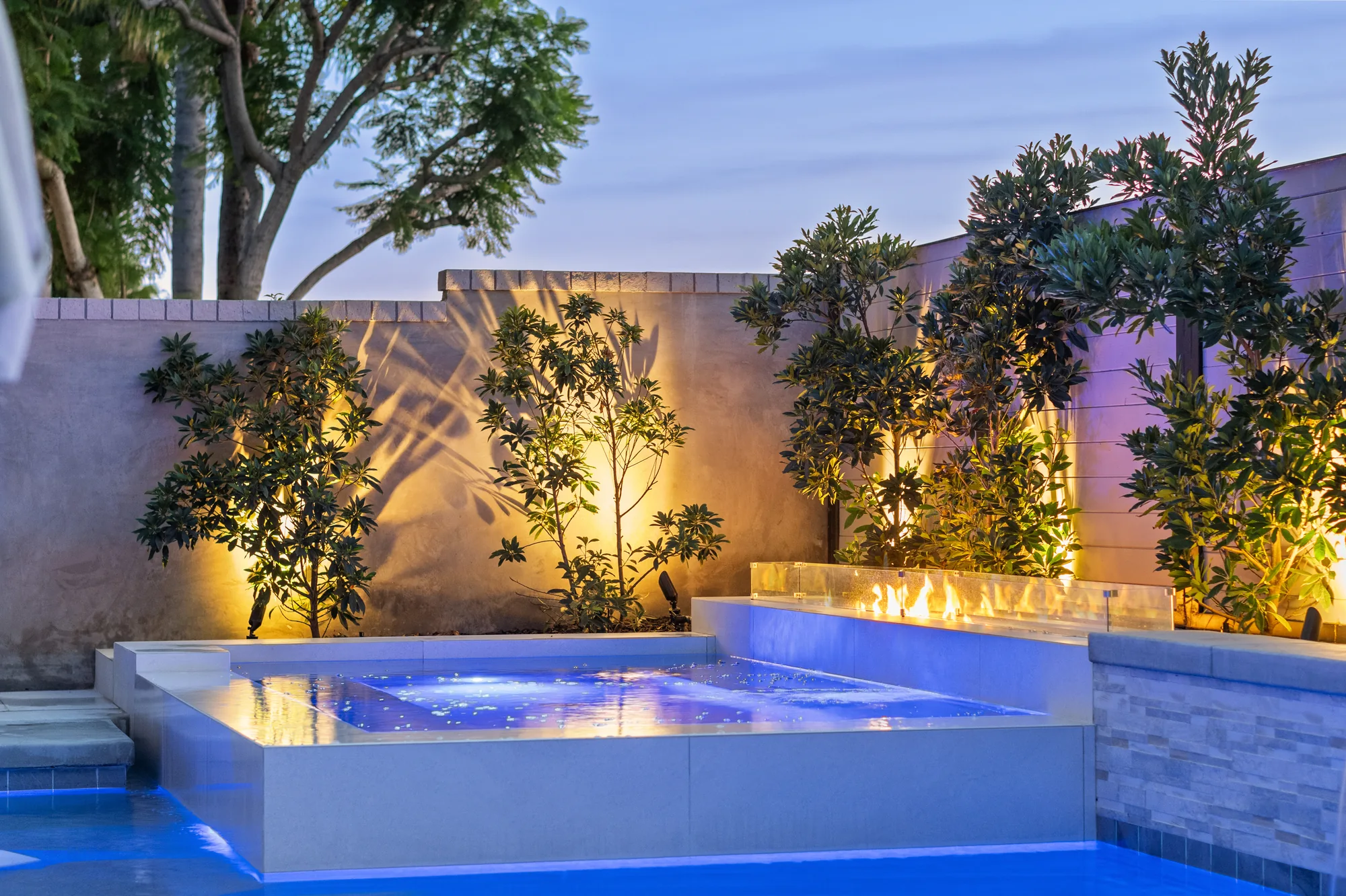Raised square spa with pale coping beside a linear fire feature and illuminated planting.