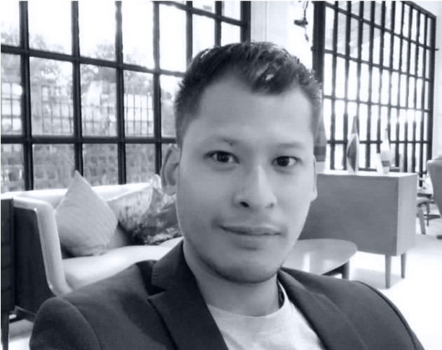 A smiling person with curly hair, wearing a leather jacket, stands in a cozy indoor setting.