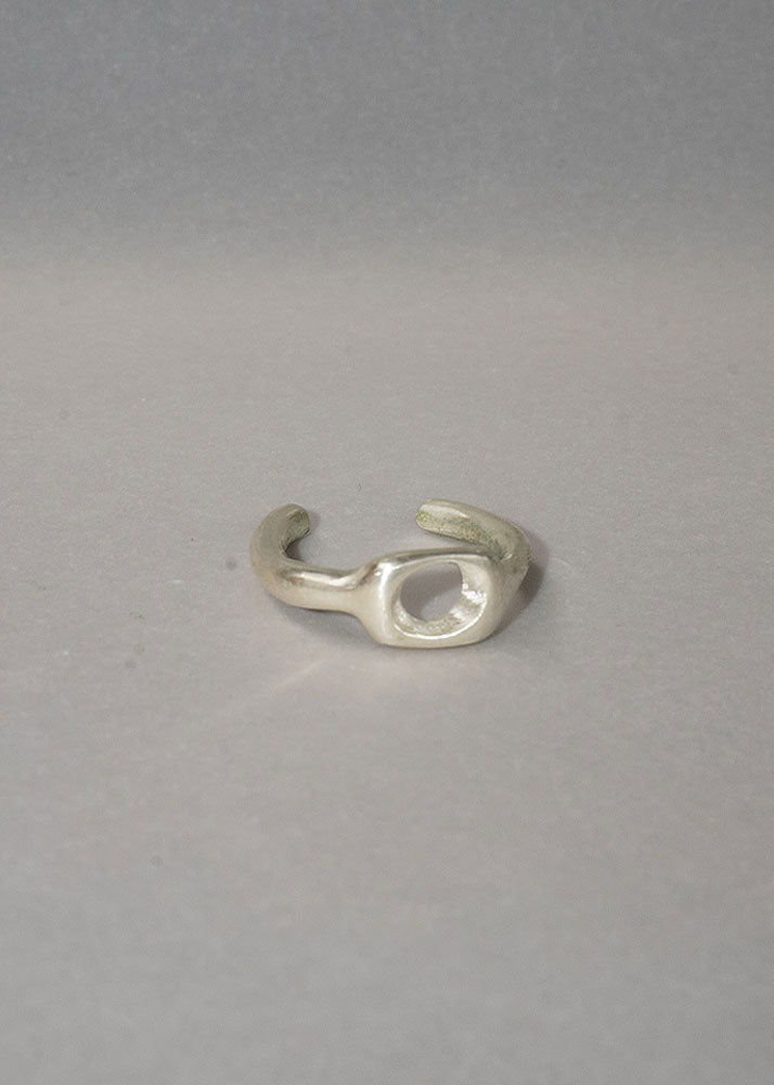 A silver abstract open ring band with a textured, organic shape, photographed on a neutral gray background.