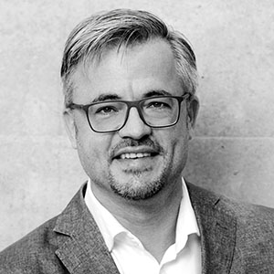 Portrait von Prof. Dr. Guido Baltes, lächelnd, mit Brille, im Sakko vor hellem Hintergrund.