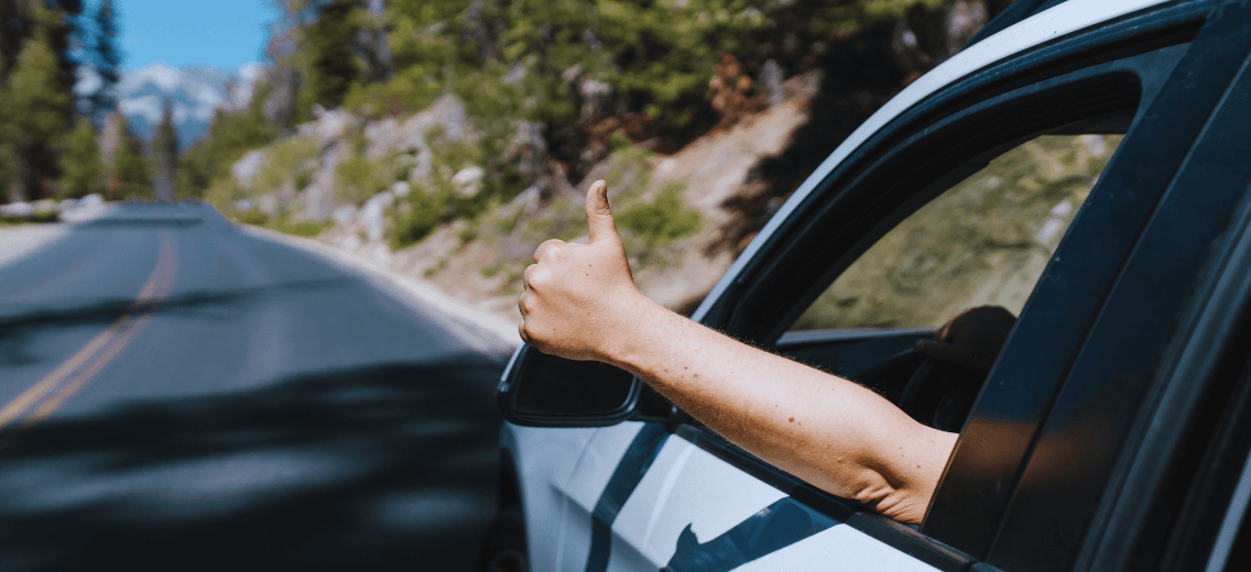 Close-up of a driver’s arm giving a thumbs-up out the car window while traveling along a scenic forest road.