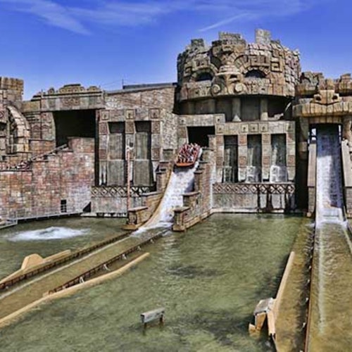 Kivettynyt vesiliukumäki, jossa on liukumäkiä ja vene, joka laskeutuu matkustajien kanssa, ympärillä kaiverruksia ja rakenteita, jotka muistuttavat raunioita.