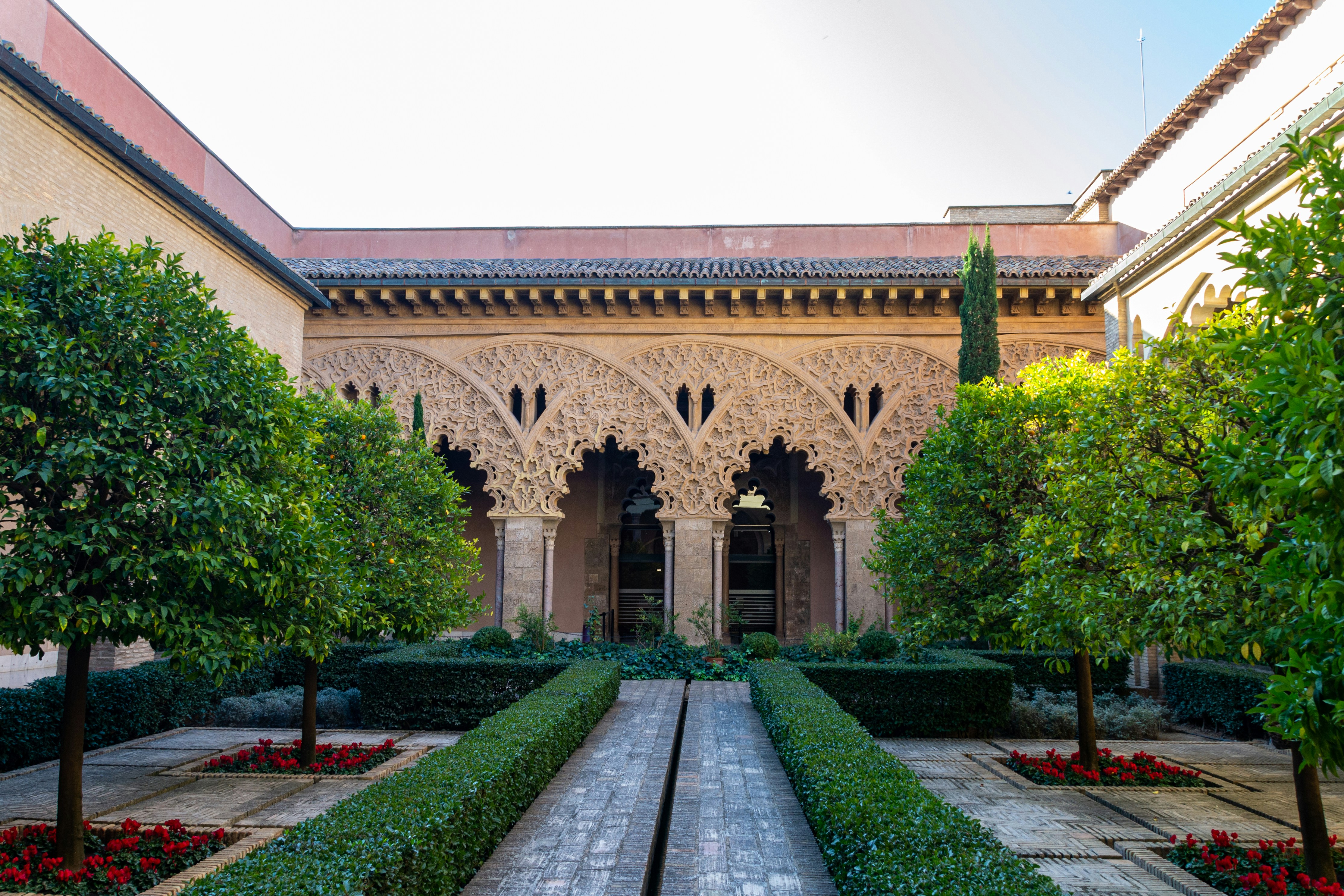 A traditional Pakistani building exterior with textured walls, framed by a lush garden, winding pathways, and scattered greenery, capturing sunlight and shadow across the courtyard.