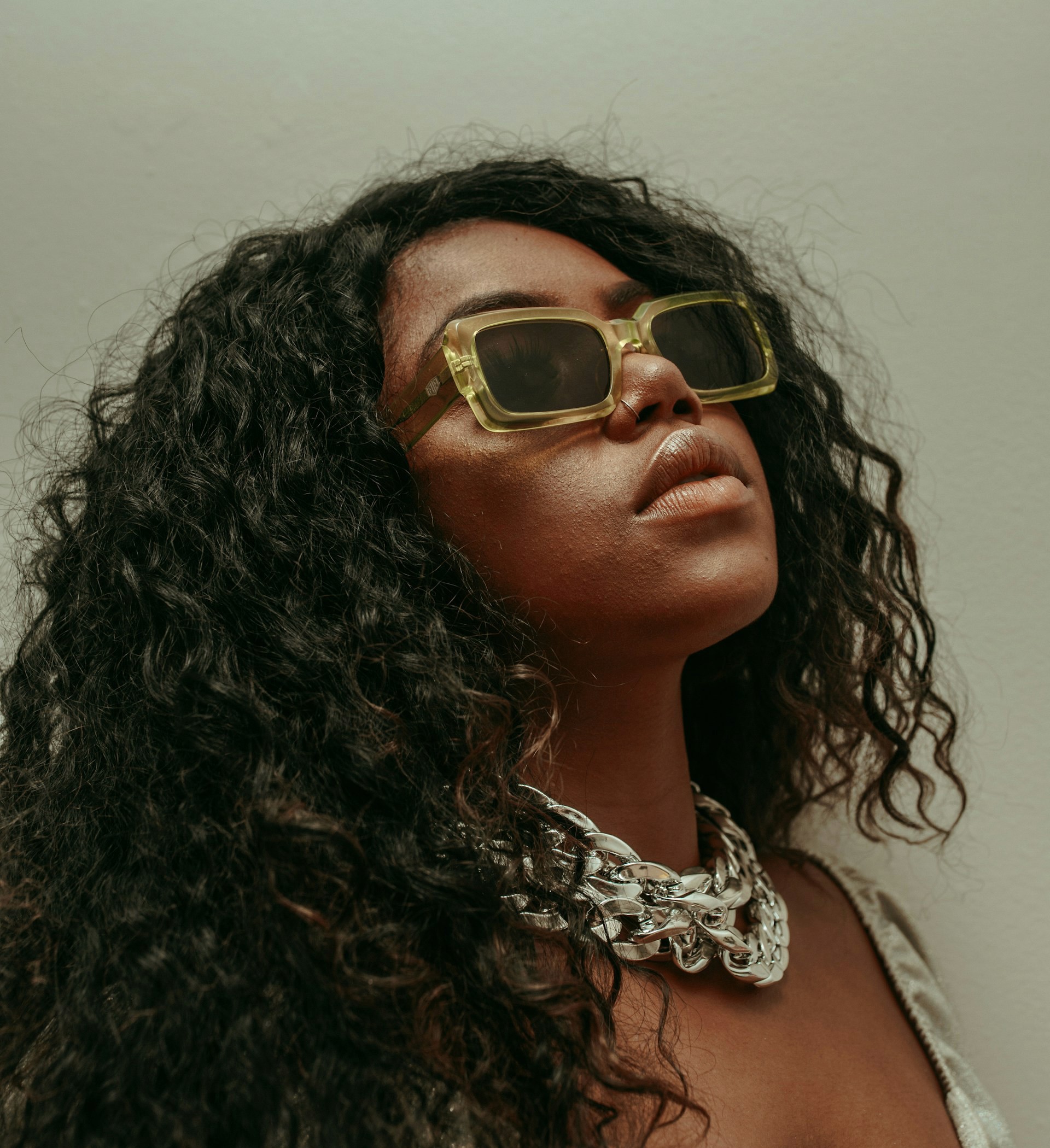 Woman with curly hair wearing yellow sunglasses and a large silver chain.