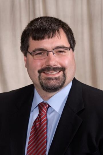 Smiling man with glasses, goatee, and suit. Wearing a blue shirt and red striped tie. Portrait photo.