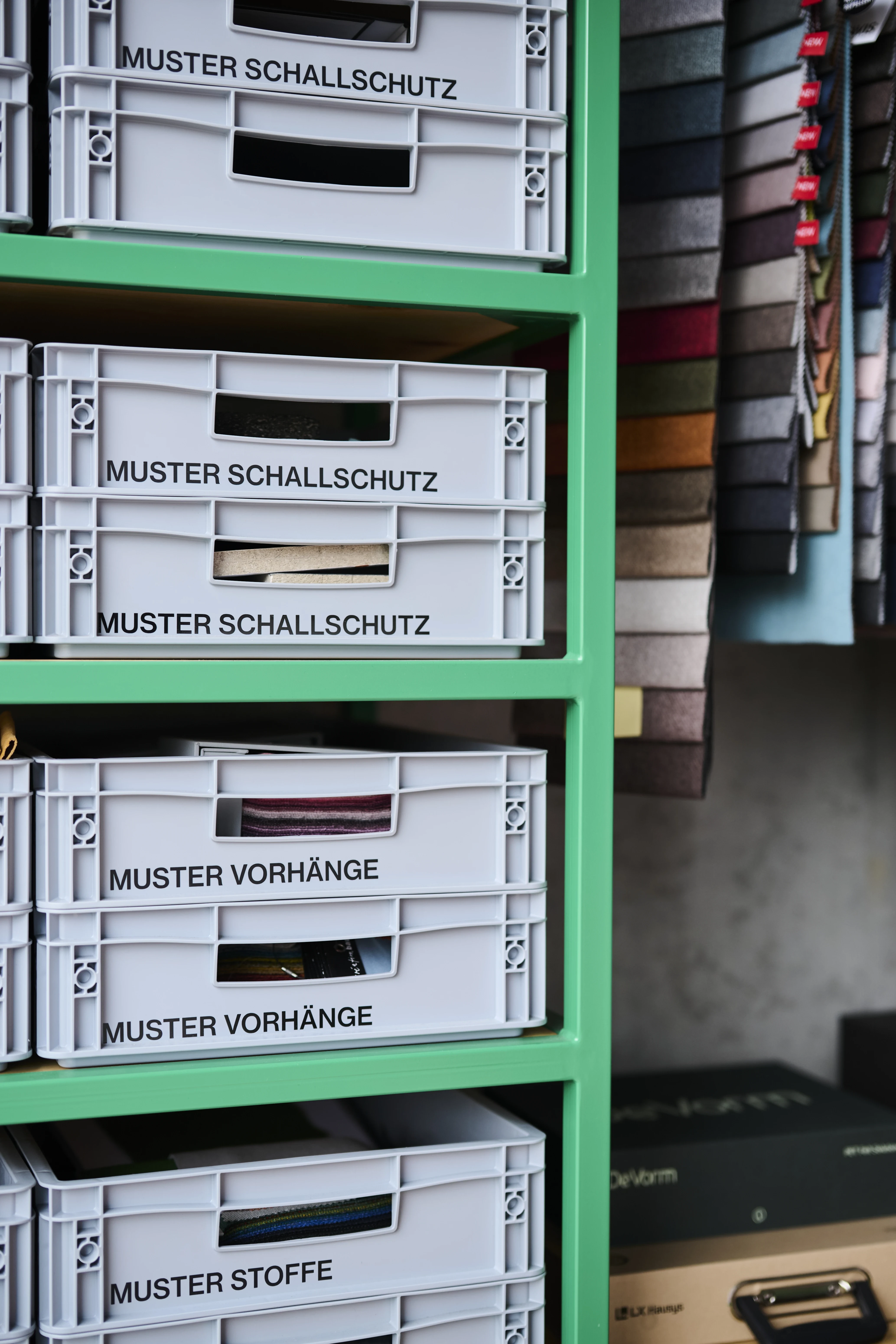 Detail of the sample archive in the UNDPLUS SHOWROOM with labelled grey crates for acoustic, curtain and fabric samples in a green shelf next to hanging textile swatches.