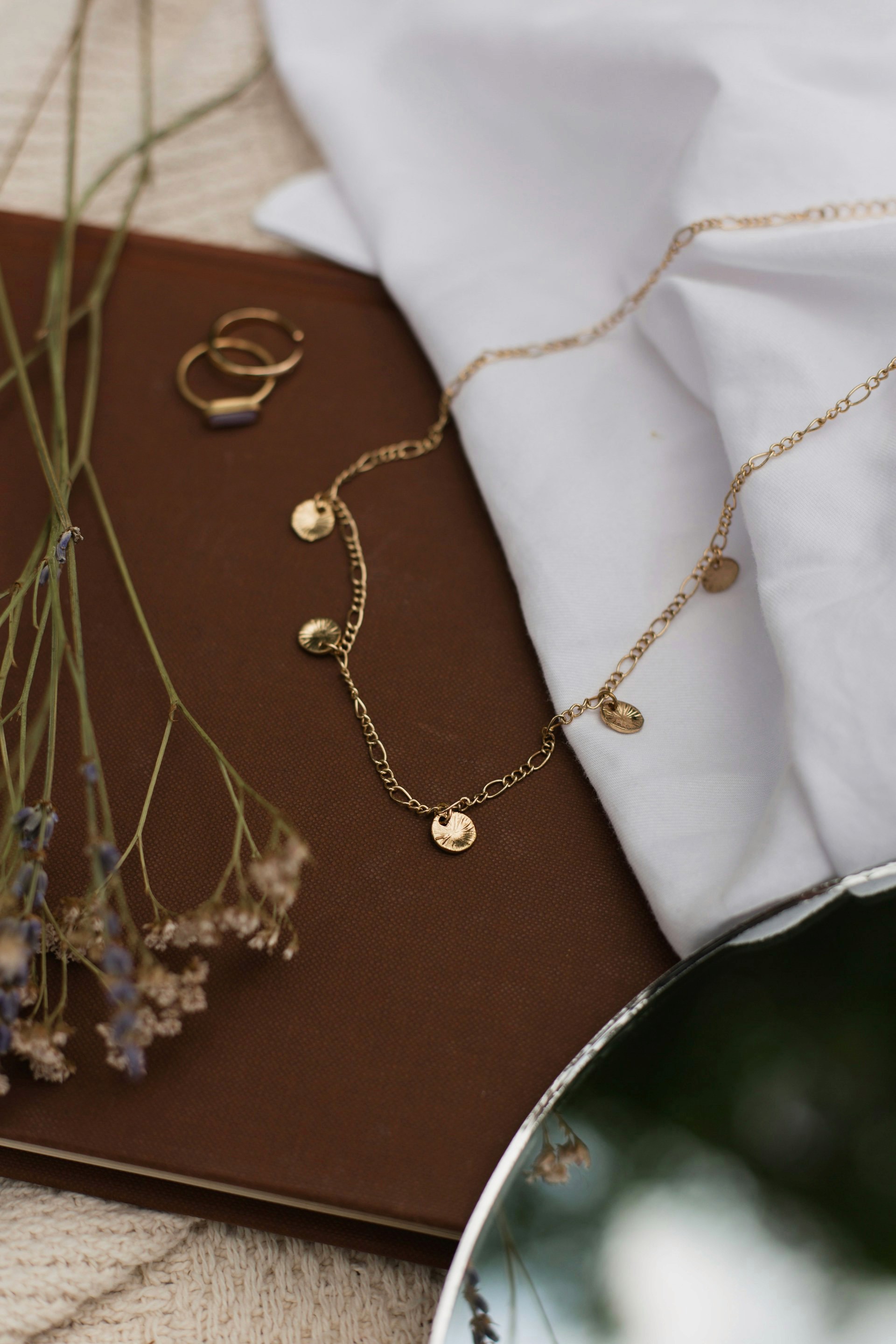 A gold charm necklace with small coin pendants laid across a brown book and white fabric.