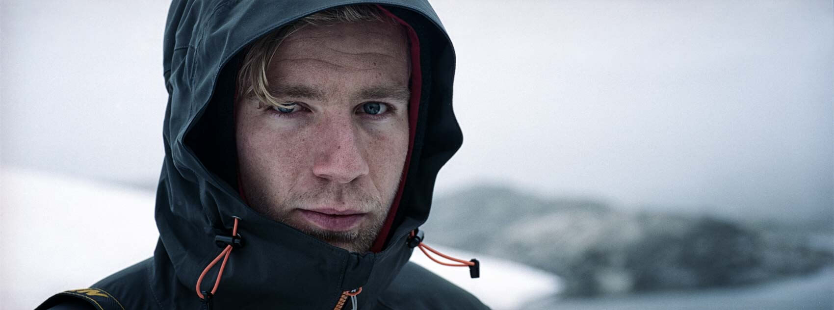 A wide-format photo capturing a hiker amidst a cold, high-altitude scene featuring snow and ice. The subject is framed against an epic background of mountains and a glacier, rendered with the soft, classic look of Portra film.