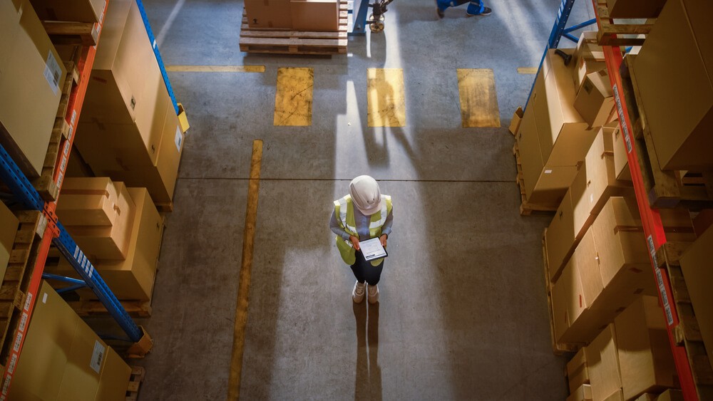 Warehouse worker in safety gear using a tablet while standing between tall shelves of stacked boxes