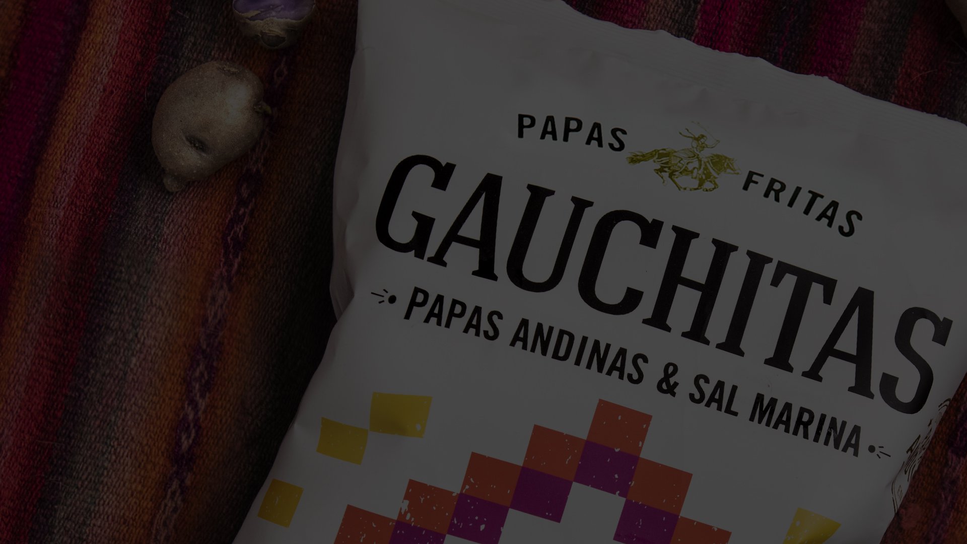 A bag of "Gauchitas Papas Fritas" chips rests on a colorful woven fabric. The packaging features geometric patterns and text highlighting Andean potatoes and sea salt flavor.
