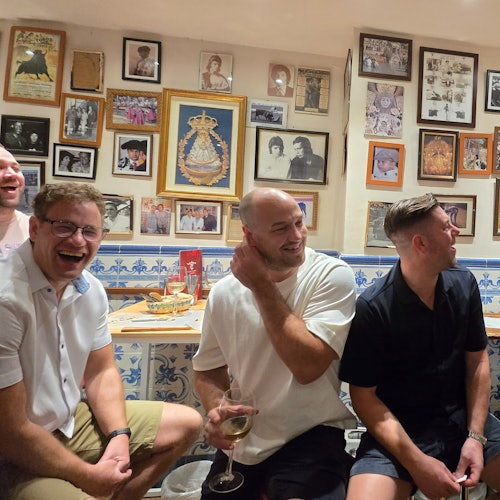 Four men sitting in a room with framed photos on the wall, laughing and chatting. One holds a glass of wine.