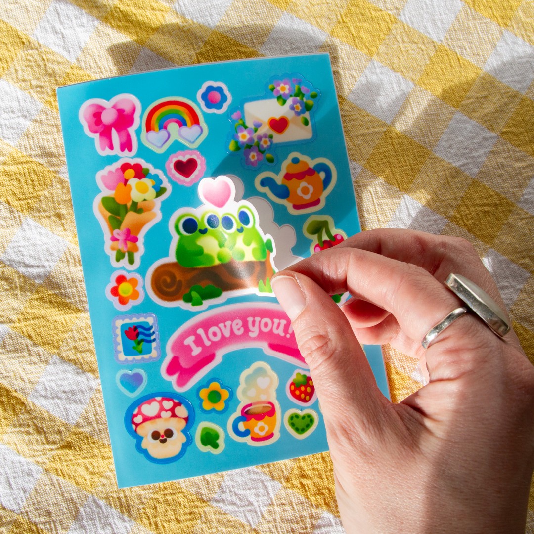 A hand peeling a sticker from a blue card. The sticker is of two cute frogs sat on a log with a heart between them. Underneath is a sticker that reads "I love you!", and there are cute stickers including a bouquet of flowers, a rainbow, and other cottagecore themed items.