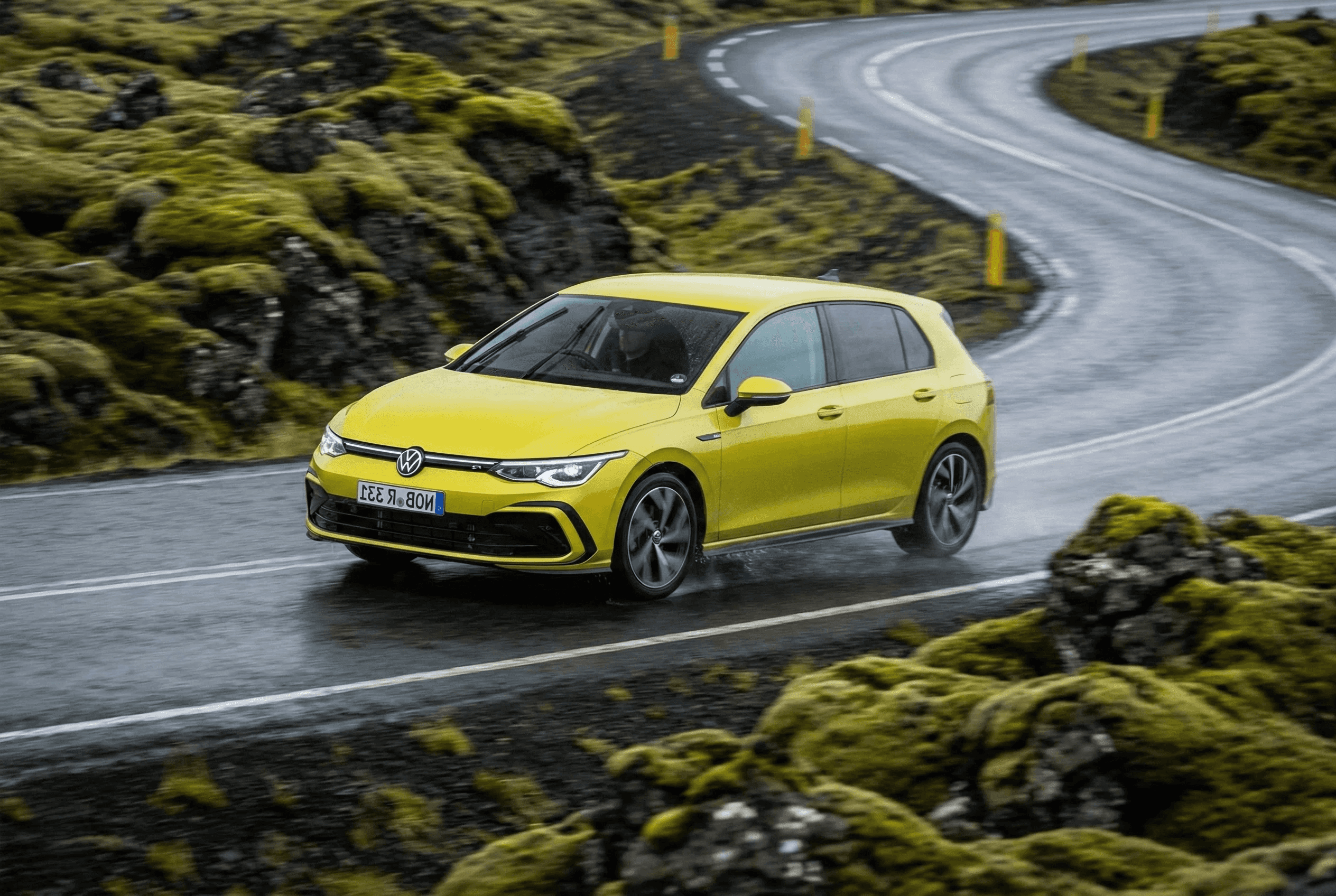 A yellow VW Golf driving on a wet asphalt road winding through a mossy lava field.