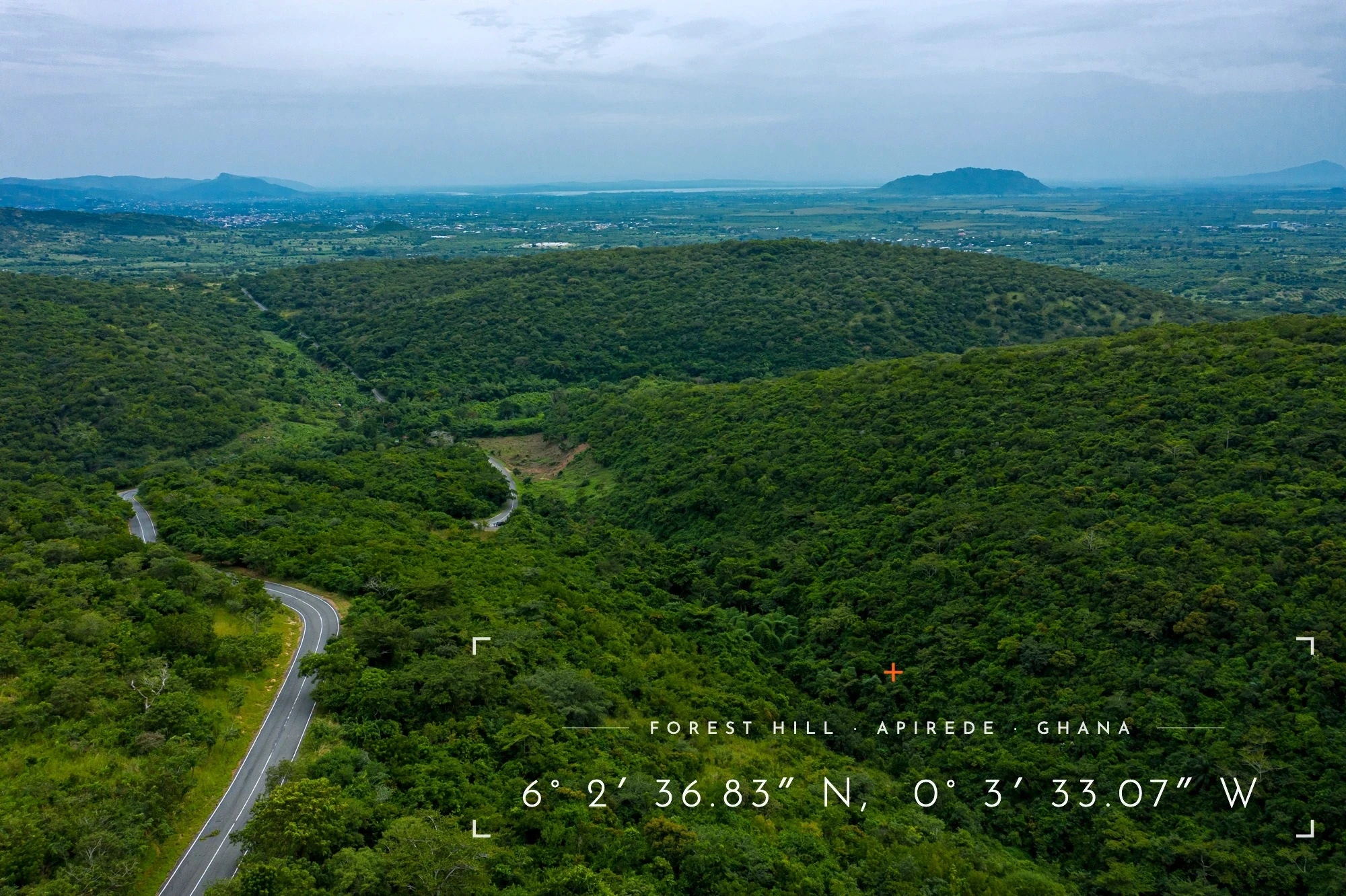 Aerial view of a winding road through forested hills with a distant valley and lake
