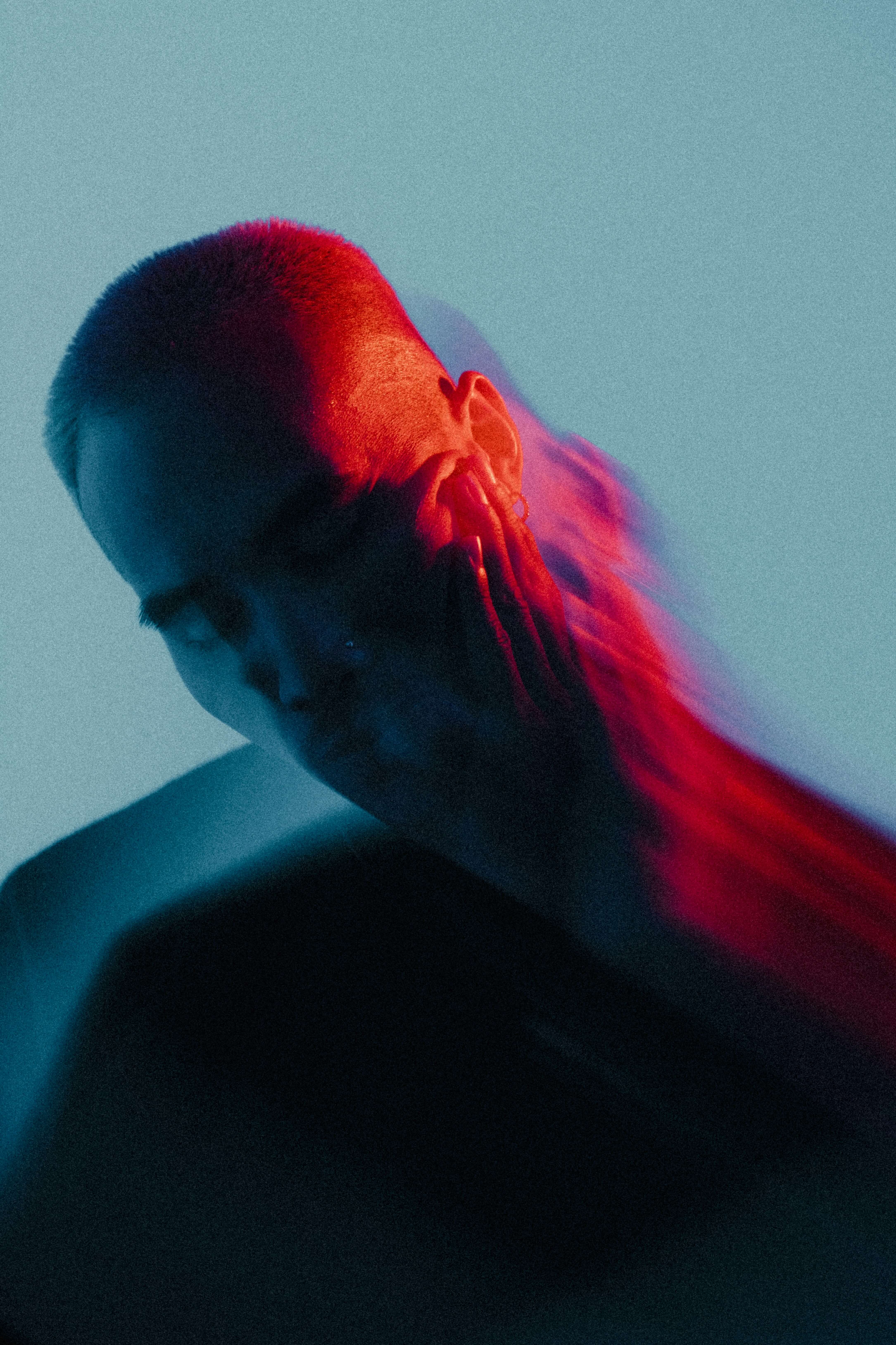 Portrait of a person with their eyes closed, hand on their face, and a blurred motion effect creating a trailing ghost-like silhouette. The subject is lit with vibrant red and blue lighting against a muted teal background, evoking a moody, introspective atmosphere.