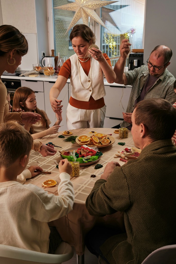 A family gathers around a table in a festive kitchen, engaging in holiday crafts with dried orange slices, beads, and string, while a large decorative star hangs in the background, creating a warm and creative holiday atmosphere.