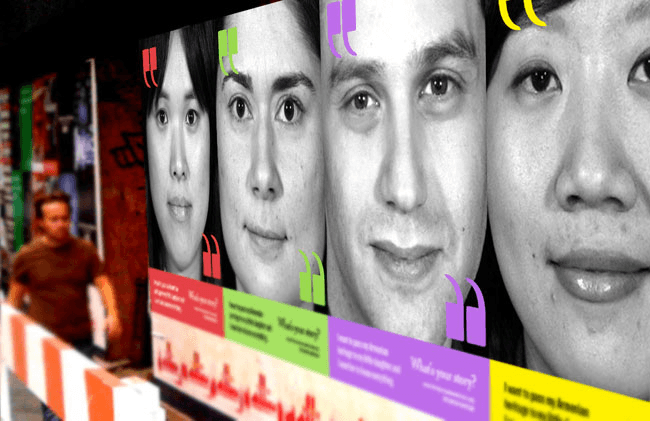 A large, colorful poster or advertisement featuring four faces—two women and two men—is displayed on a wall. The faces are arranged side by side, with each person shown in a close-up view. The poster includes text in red, green, and purple, arranged in horizontal panels below the faces. A blurred figure of a person in a brown shirt is visible to the left of the poster. The image is primarily in black and white, with the colorful elements providing contrast.