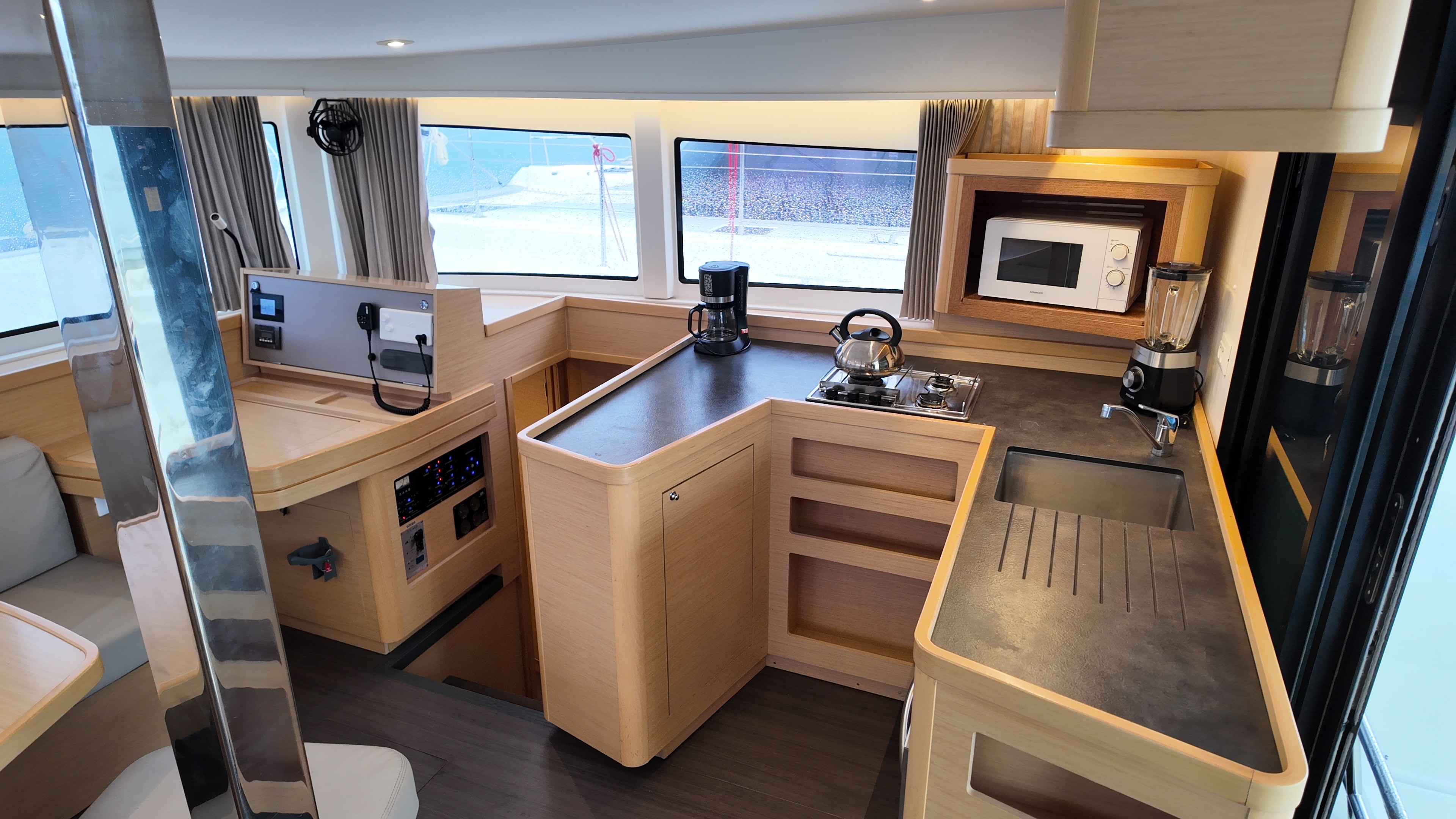 Lagoon 42 catamaran saloon and galley interior showing the full U-shaped galley layout with light oak cabinetry, dark worktops, two-burner hob, filter coffee machine, microwave, blender, stainless sink, inverter panel, and panoramic saloon windows looking out over the marina