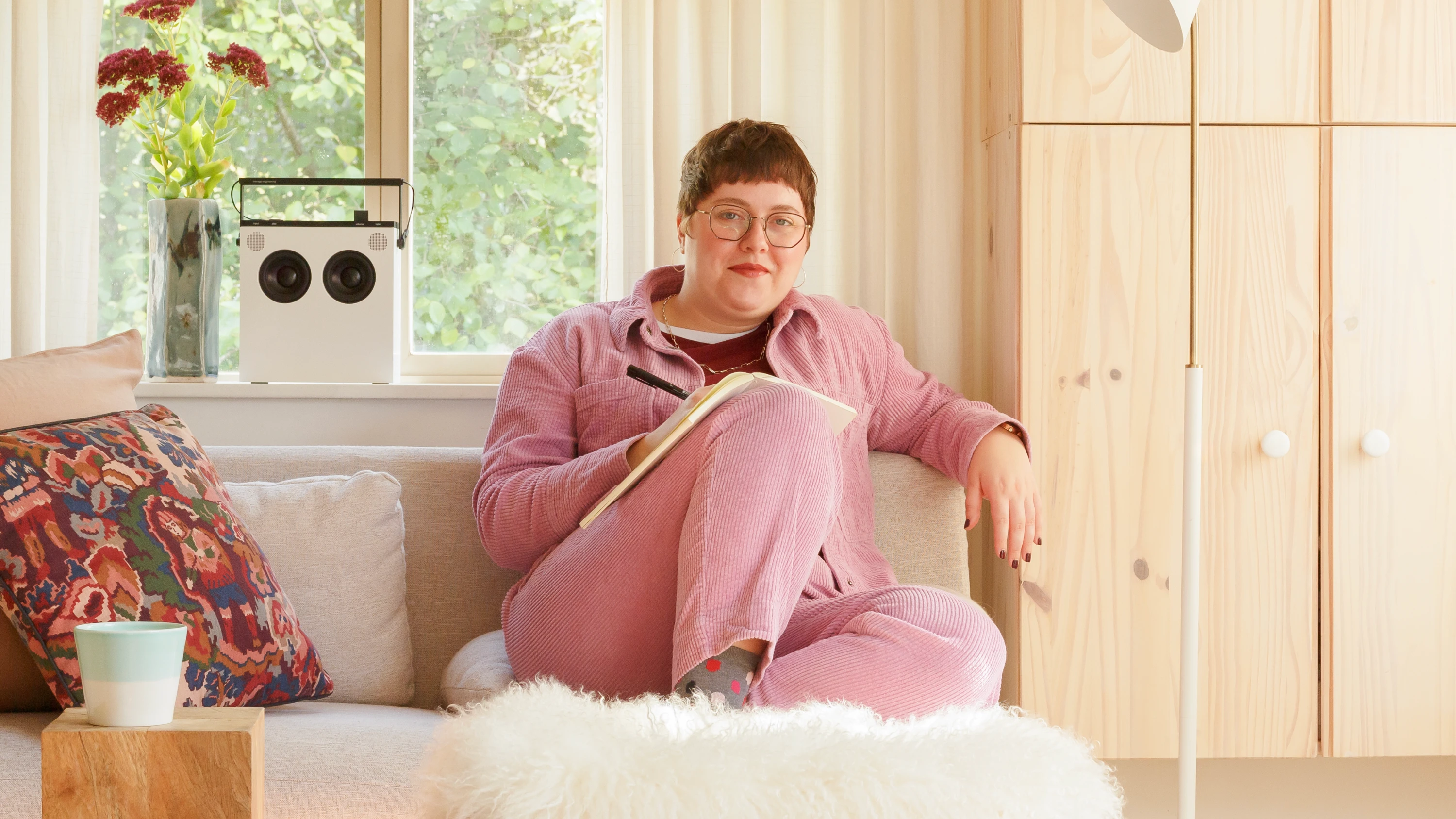 Emily Kiana sitting on a couch wearing a pink corduroy suit holding a sketch book and pen.