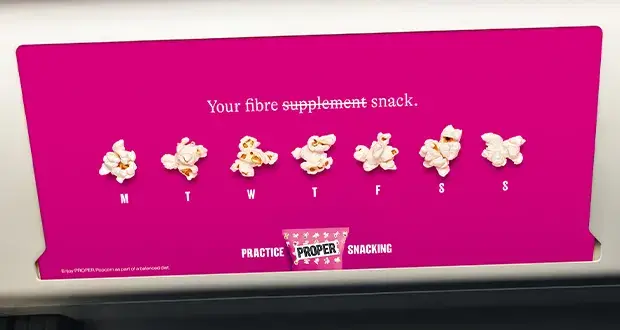 Pink PROPER Snacks advert from a creative campaign on fibremaxxing, showing pieces of popcorn arranged across the days of the week with the line “Your fibre supplement snack.”