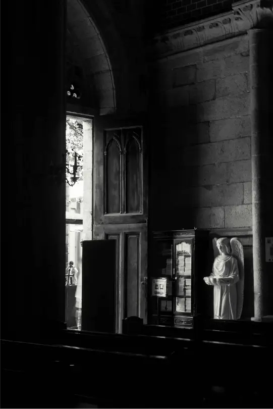 Black and white interior of a stone church, featuring a white angel statue holding a basin next to a wooden cabinet, with light streaming in from an open door.