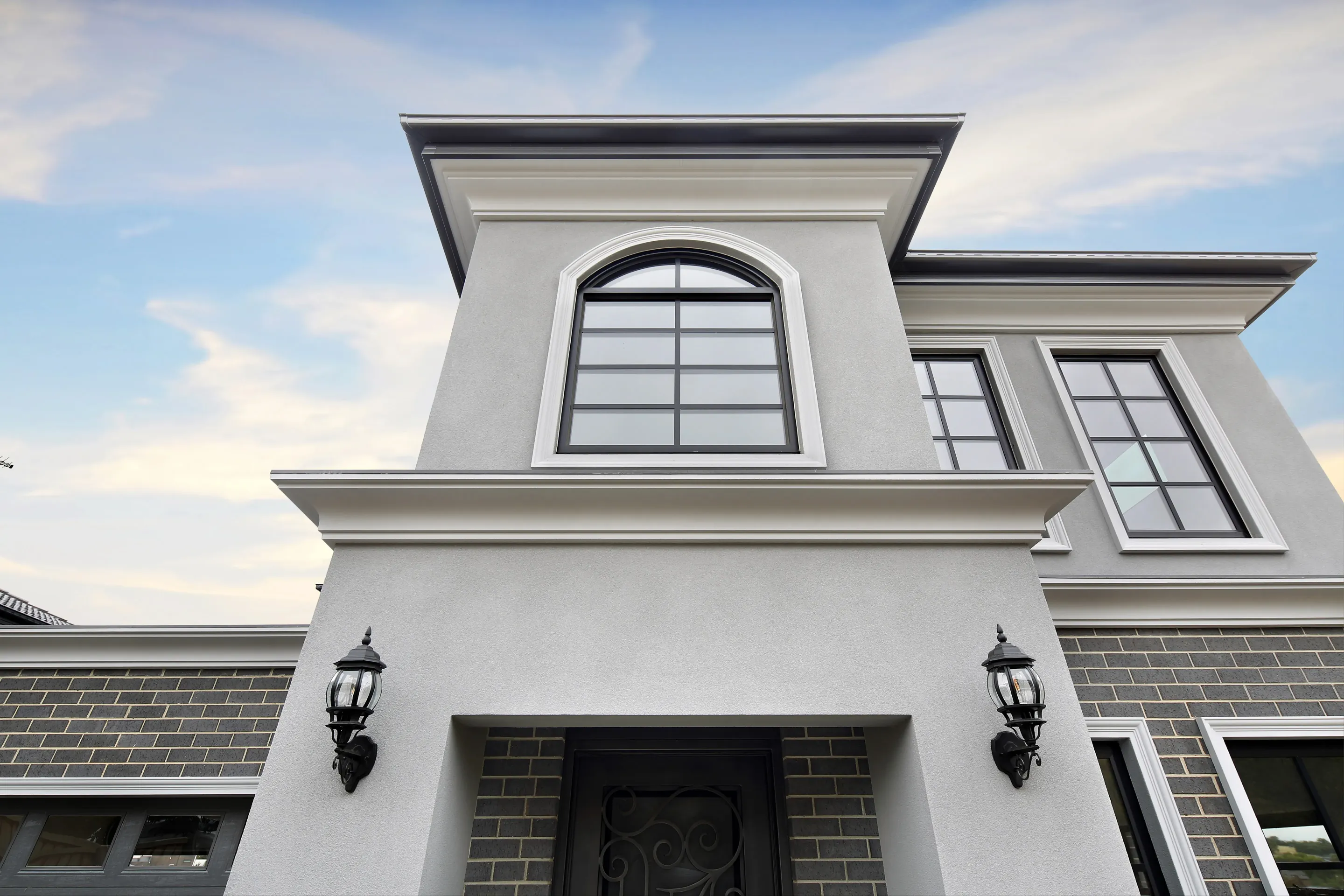 Entry portico of South Morang Residence seen from below, with grey render, moulded cornices, an arched feature window, face brick, carriage lanterns and a wrought-iron and glass entry door
