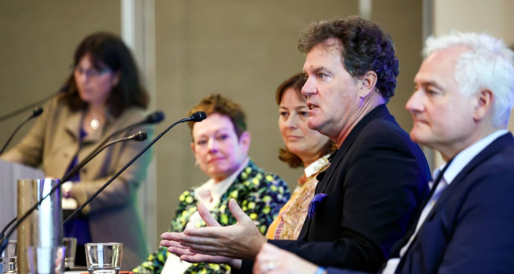 Panel of speakers at an event, with a woman standing and holding a microphone, discussing various topics.