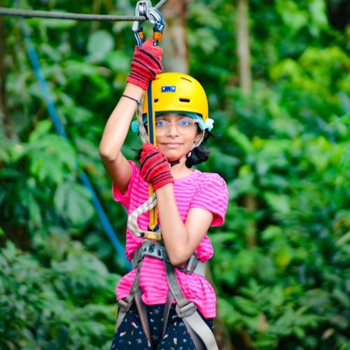 一个戴着黄色头盔、穿着粉红色衬衫的孩子在郁郁葱葱的绿色森林中抓住了拉链的安全带。
