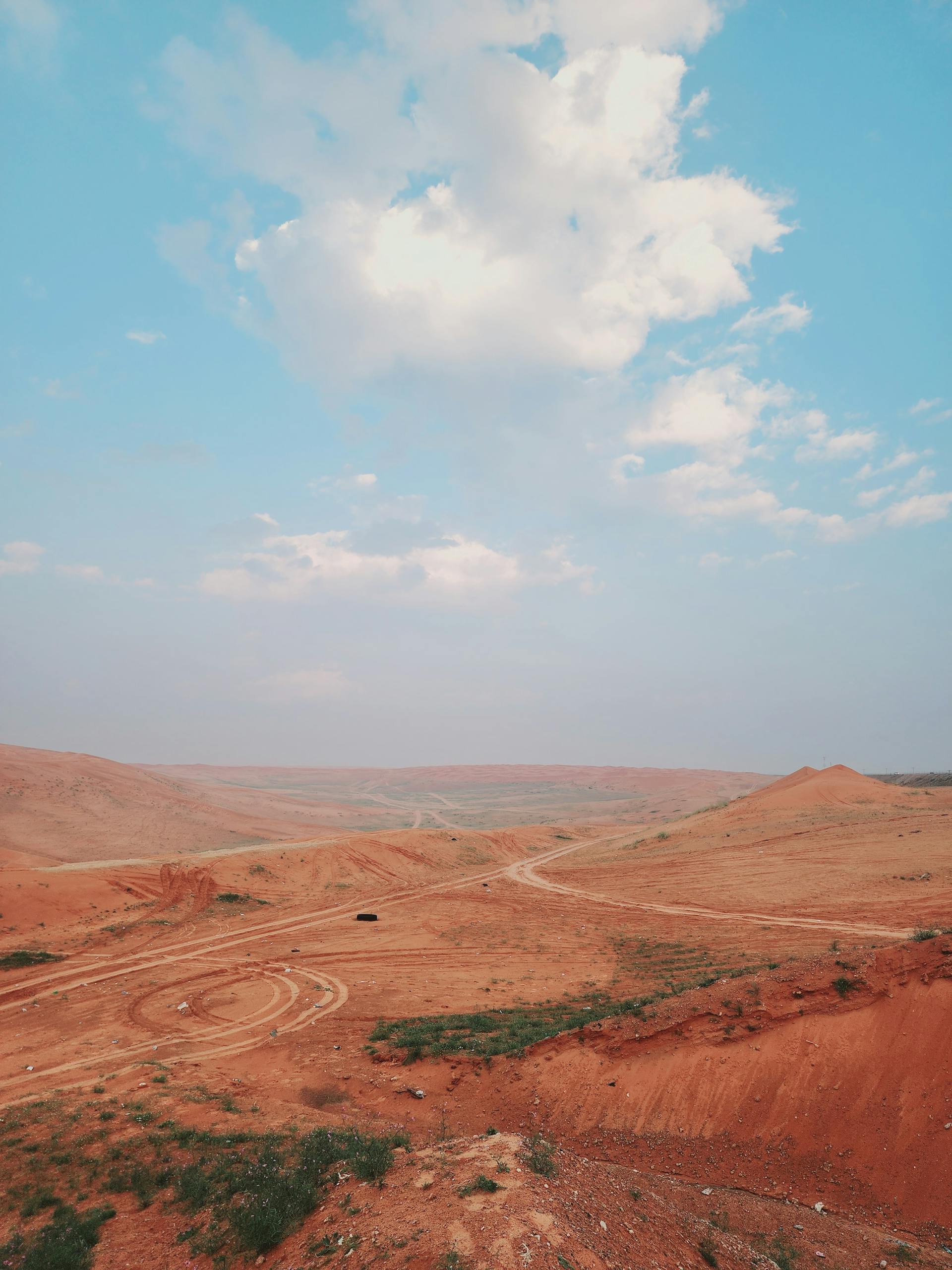 Desert landscape with dirt roads under a blue cloudy sky. The terrain is reddish-brown, with green accents.