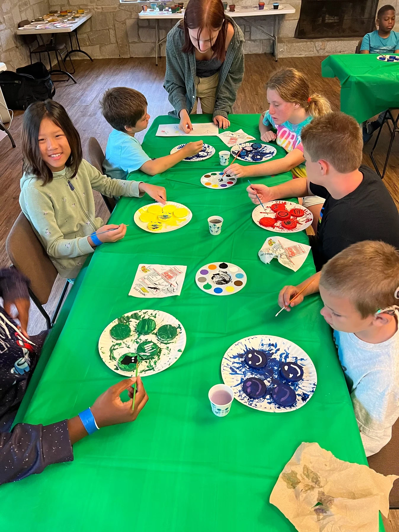 A group of campers and staff participate in a painting activity. They are seated around a green table with paint palettes and paint supplies.