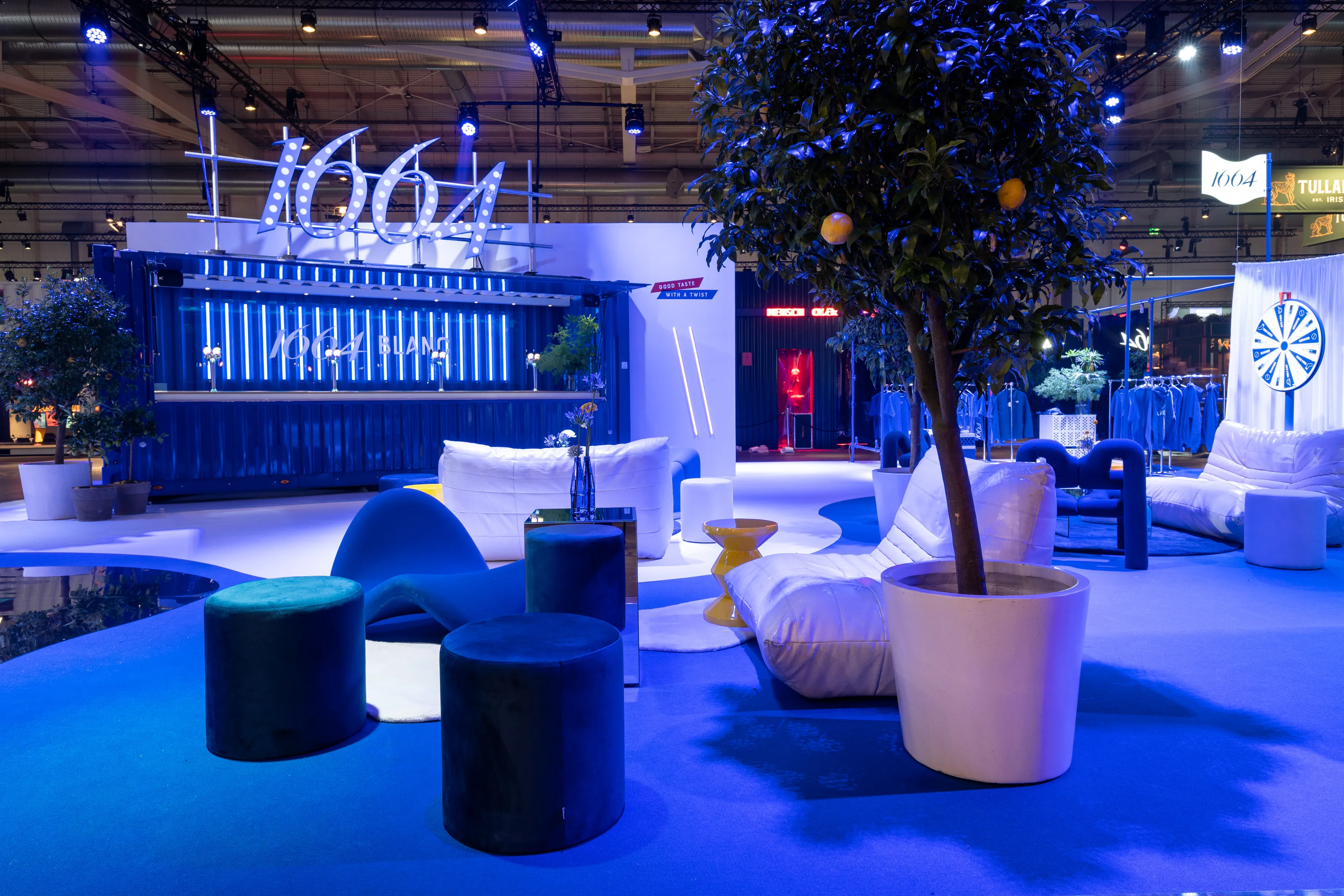 Blue-lit lounge of the 1664 Blanc booth with white beanbag sofas, velvet poufs and citrus tree in front of the container bar.