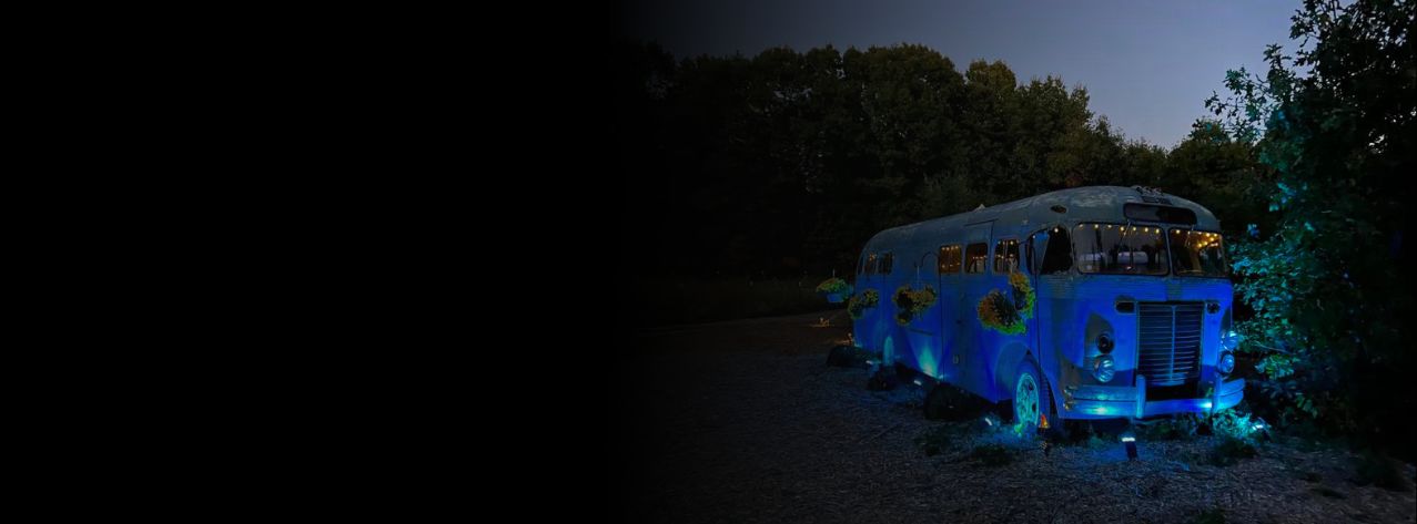 A vintage bus lit up by blue lights outside in the dark with flowers hanging off the side