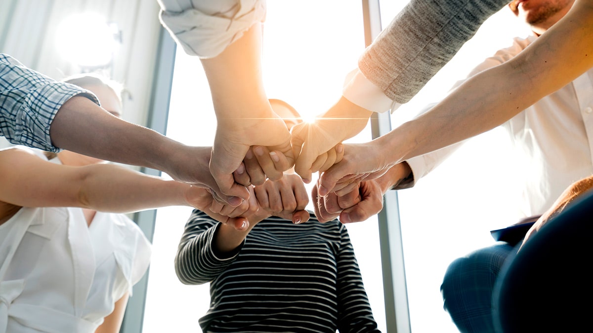 Hands of diverse individuals join together in a supportive gesture, with light shining brightly in the background.