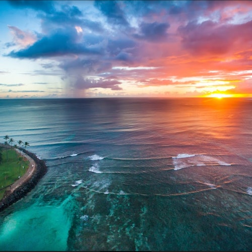 วิวมุมสูงของมหาสมุทรในยามพระอาทิตย์ตกที่มีท้องฟ้าสีส้มและสีม่วงที่สดใส, คลื่นสงบ, แนวชายฝั่งที่มีต้นปาล์ม, และเมฆระเบิด.