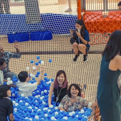 Adults laughing and playing in a ball pit filled with blue and white balls, while a woman takes photos from above.