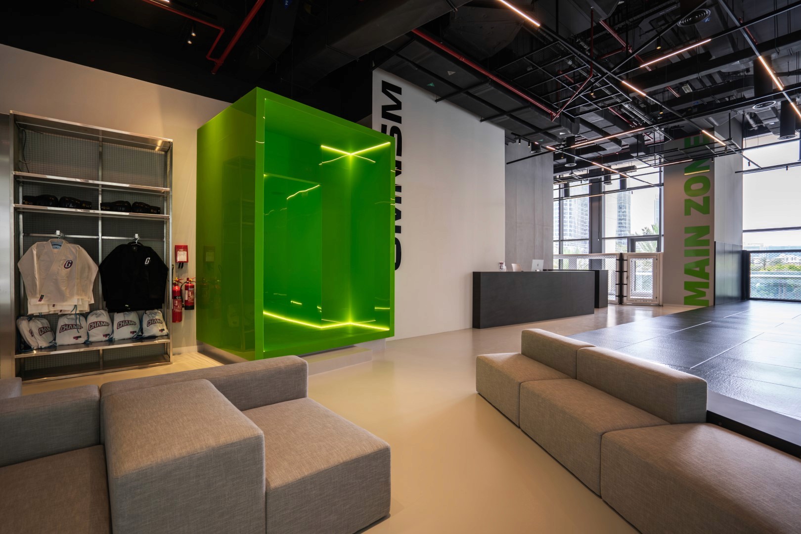 Modern gym lobby featuring a sleek design with gray sofas, a bright green glass meeting room, and shelves displaying merchandise, against a backdrop of industrial-style ceilings and wall decor.
