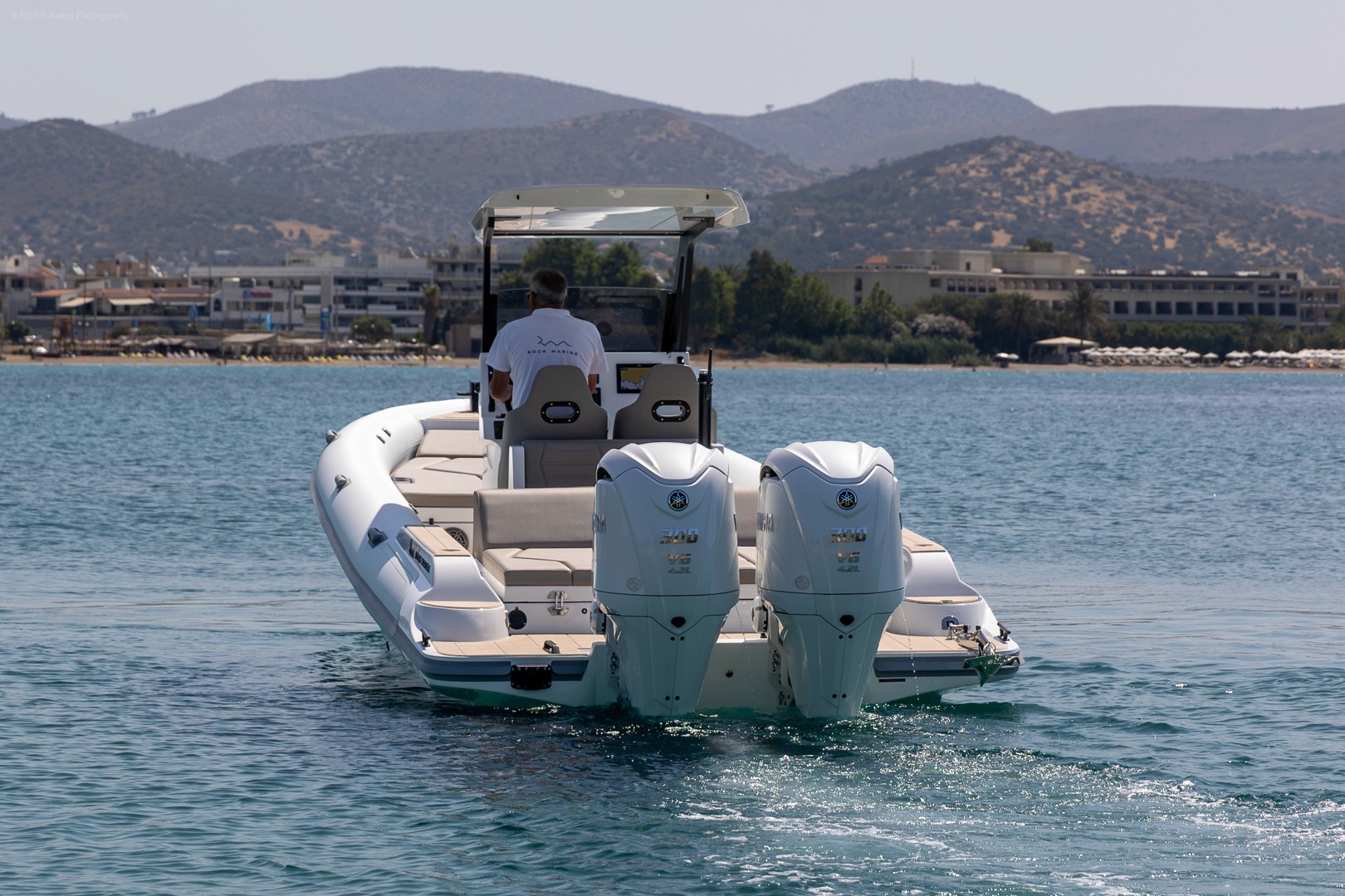 White inflatable speedboat with twin outboard engines cruising in calm turquoise waters near Greek coastal hills and waterfront buildings.